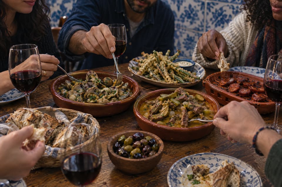 Amigos a partilhar amêijoas, pica-pau, peixinhos da horta, chouriço e azeitonas numa mesa de madeira, com copos de vinho tinto.